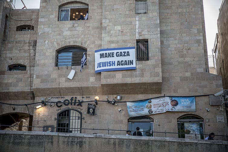 A blue-and-white sign hangs from a window on a three-story stone building.
