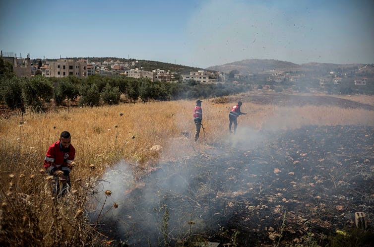 Three people walk through a field where the remains of a fire are smoldering.