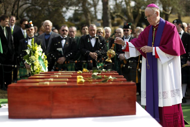 Man wearing red, purple and white vestments shown incensing caskets as crowd of people look on