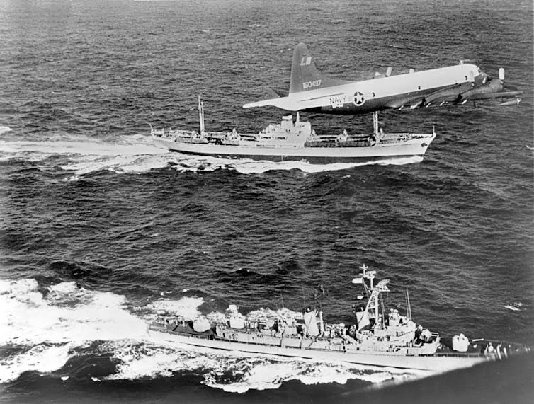 Two ships steam side by side with an aircraft flying overhead.