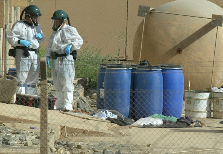 Two people in protective clothing, helmets and masks stand near blue barrels outside a building.