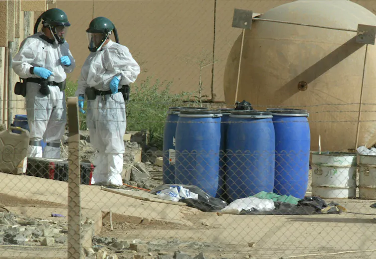 Two people in protective clothing, helmets and masks stand near blue barrels outside a building.