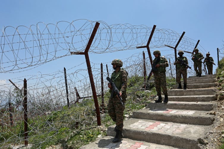 Armed soldiers walk next to a barbed-wire fence.