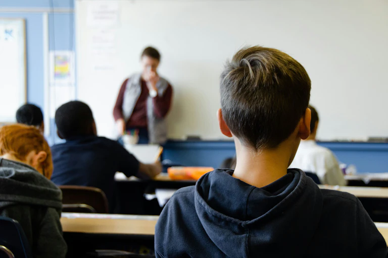 a boy seen from behind sitting in a classroom. Other students and a teacher are also seen in the background
