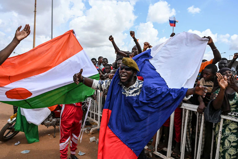 Man wearing military fatigues is draped with a white, blue and red flag while a crowd behind him holds their hands up; there is an orange, white and green flag off to the side