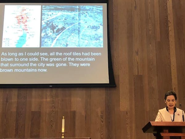 A person stands at a lectern in front of a screen with photos and text reading 'As long as I could see, all the roof tiles had been blown to one side. The green of the mountain that surround the city was gone. They were brown mountains now.'