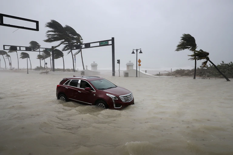 An SUV is stuck in rising flood water as the ocean pours in. Palm trees blow in the hurricane winds.