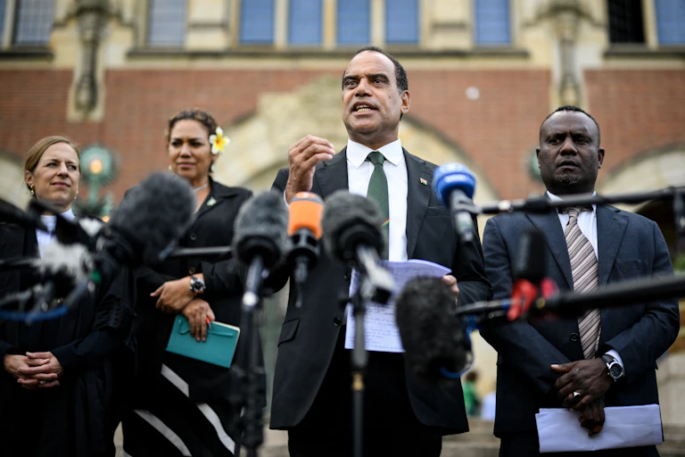 A man gestures as he speaks before a bank of media microphones. A woman with a tropical flower in her hair is behind him.