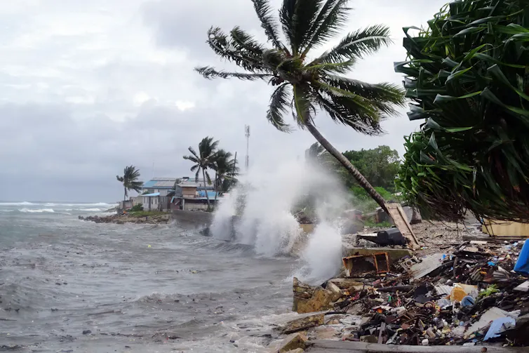 Waves spend spray higher than houses and lap at the edges of homes, with palm trees in the background.