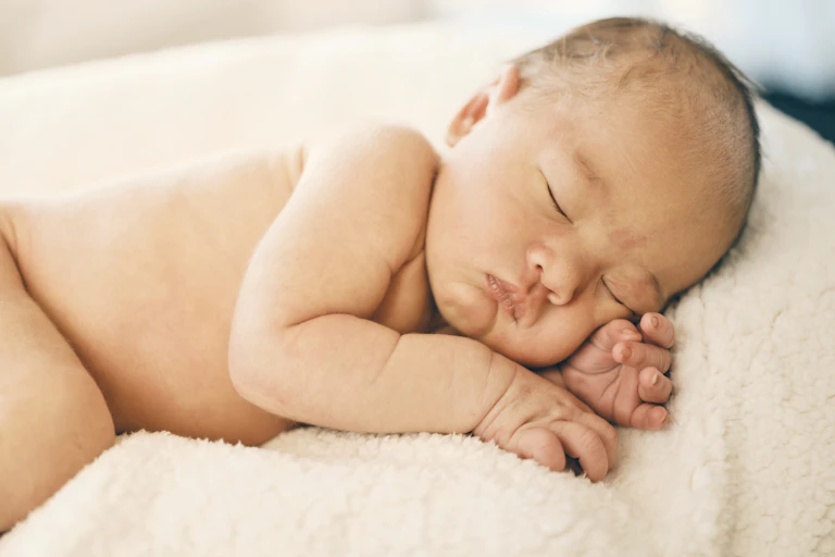 A naked newborn baby sleeping on its side on a soft off-white blanket