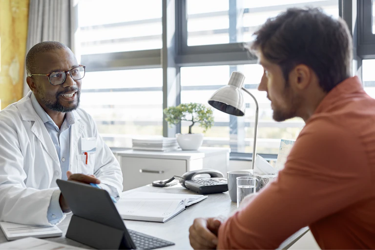A doctor in a lab coat speaks with a young man wearing casual clothes. They are sitting at a desk.