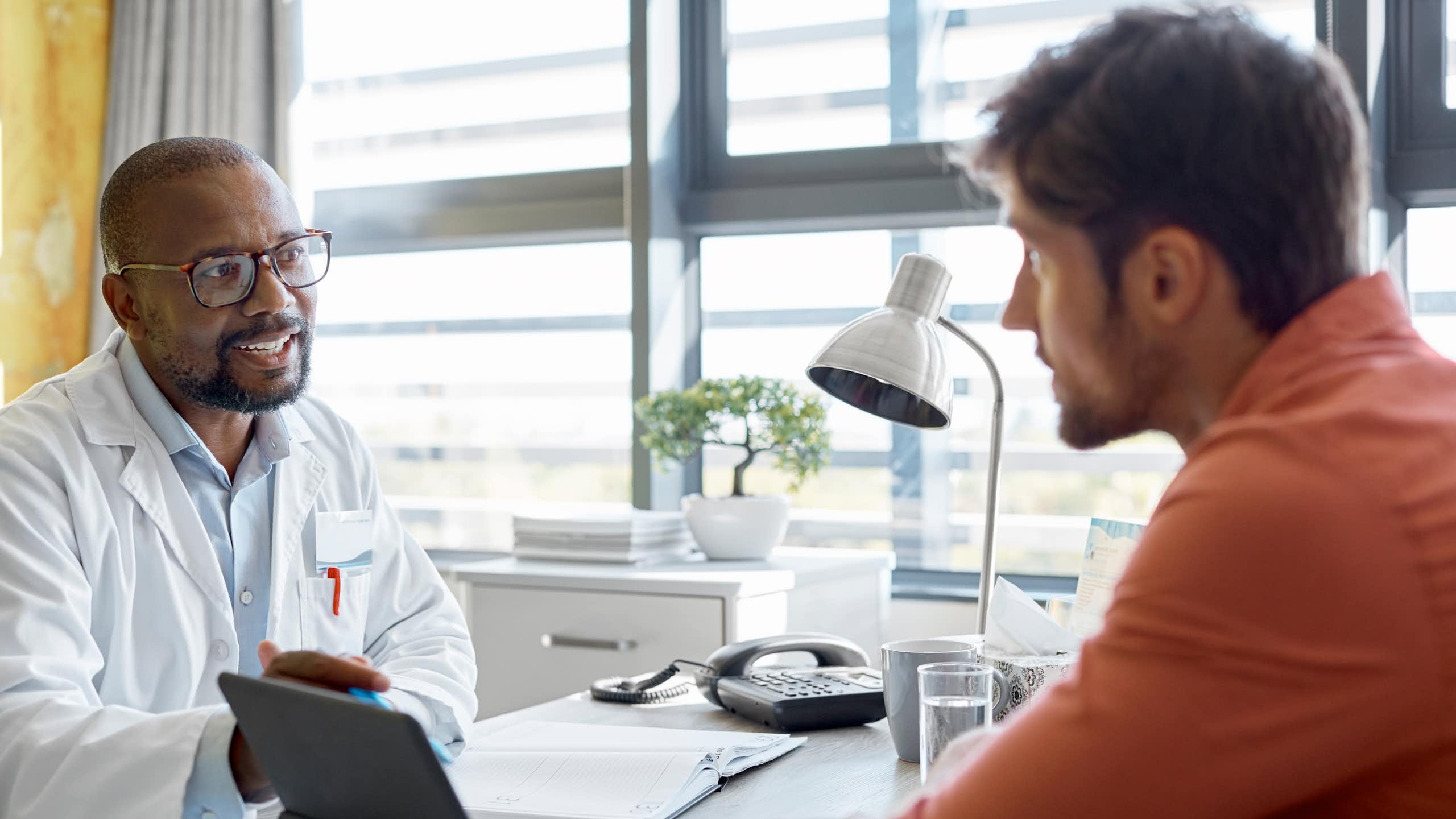 A doctor in a lab coat speaks with a young man wearing casual clothes. They are sitting at a desk.