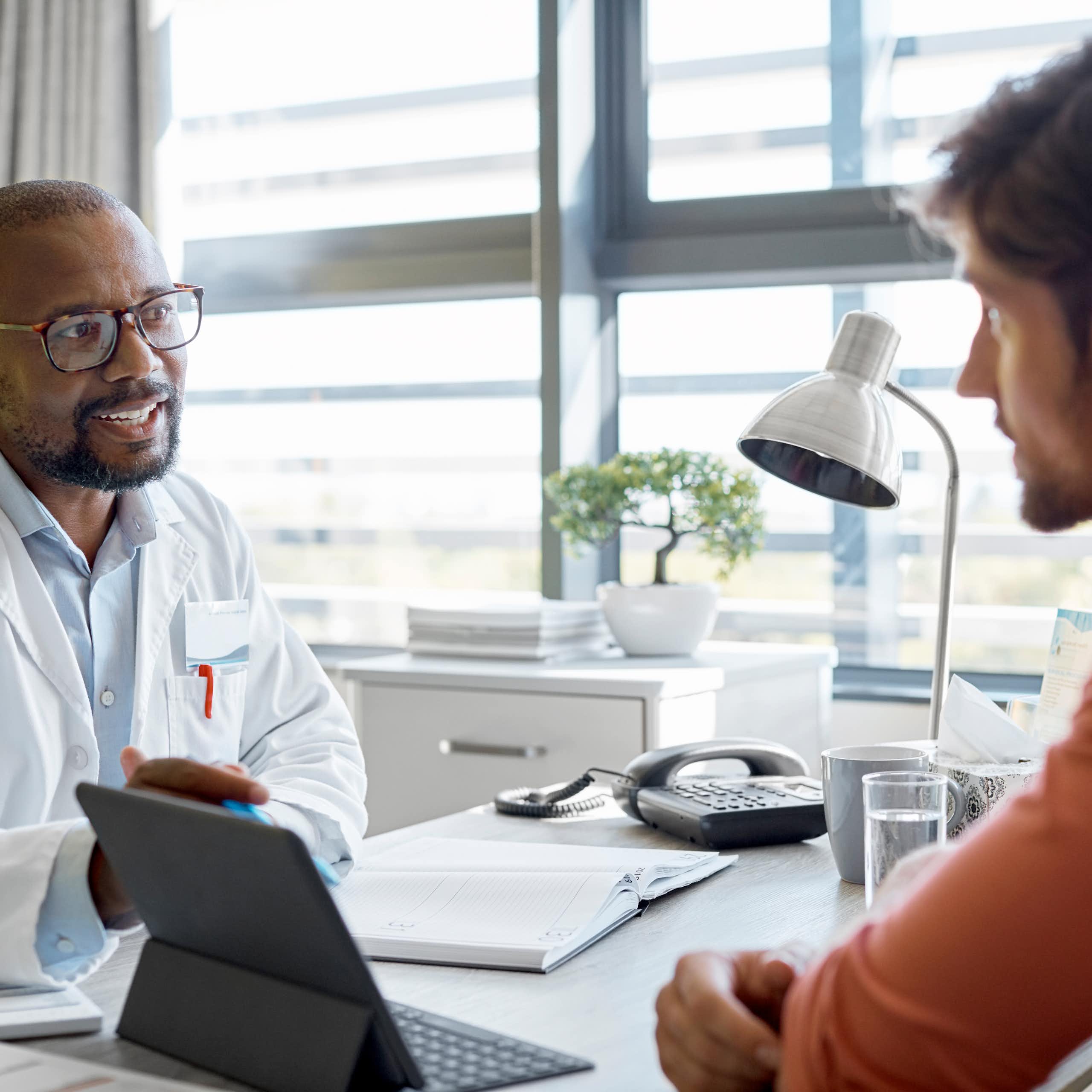 A doctor in a lab coat speaks with a young man wearing casual clothes. They are sitting at a desk.