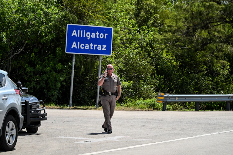A law enforcement officer talks on his phone as he walks by a bright blue sign reading "Alligator Alcatraz."