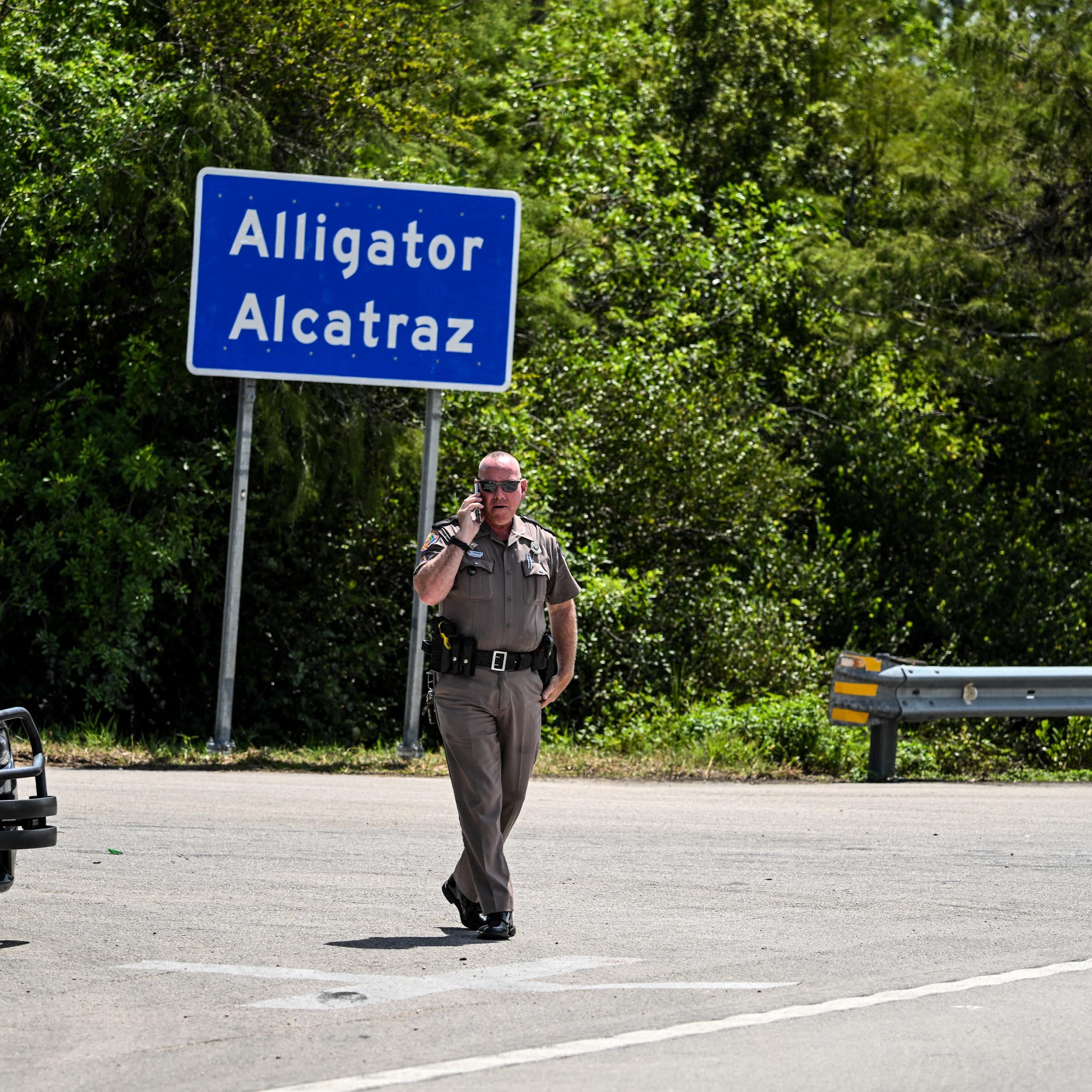 A law enforcement officer talks on his phone as he walks by a bright blue sign reading "Alligator Alcatraz."