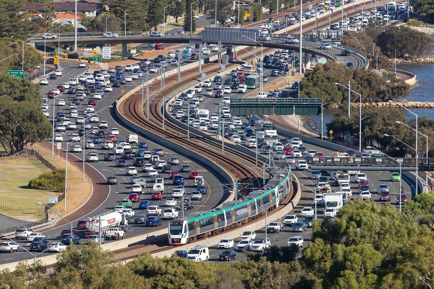 Aerial view of heavy traffic on a freeway in Perth, Australia, with a train running along the parallel rail line, multiple lanes congested with vehicles.