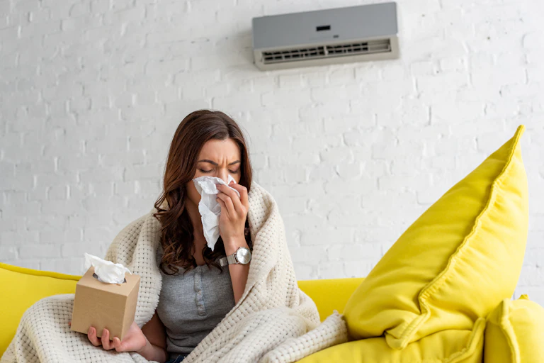 A woman sits on a couch in front of an air conditioning unit wrapped in a blanket. She blows her nose into a tissue.