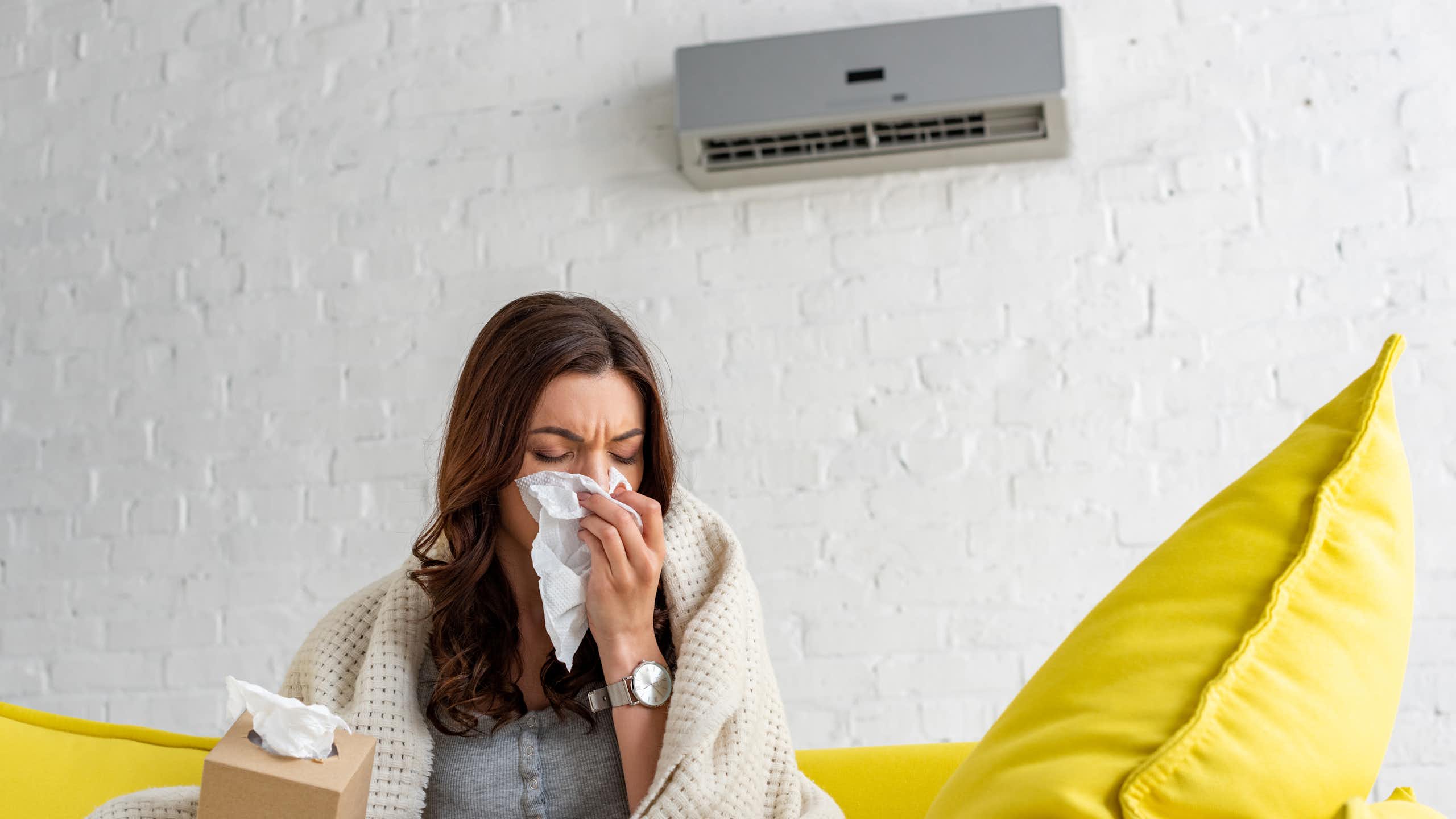 A woman sits on a couch in front of an air conditioning unit wrapped in a blanket. She blows her nose into a tissue.