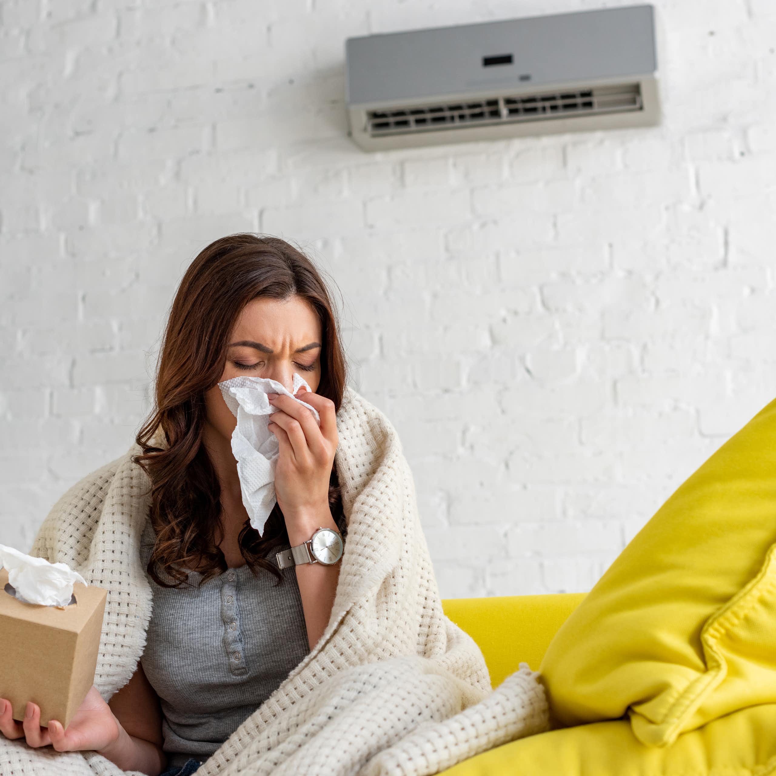 A woman sits on a couch in front of an air conditioning unit wrapped in a blanket. She blows her nose into a tissue.
