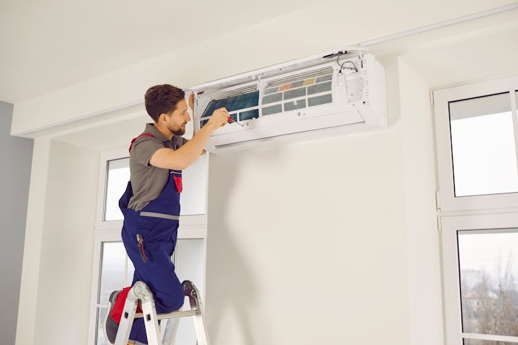 A man standing on a ladder repairs an air conditioning unit that's mounted to the wall.