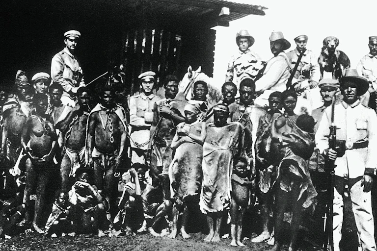 A very old black and white photo shows nearly naked Africans chained together with uniformed soldiers posing among them.