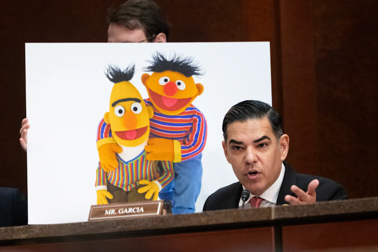A person holds a large photo of Bert and Ernie, the 'Sesame Street muppets,' behind a man in a suit and tie who is speaking.