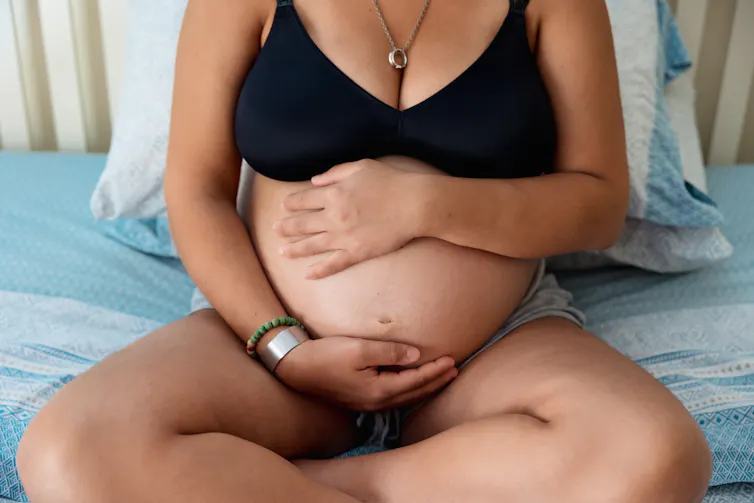 Close up view of a pregnant woman touching and holding her belly while sitting on the bed. -