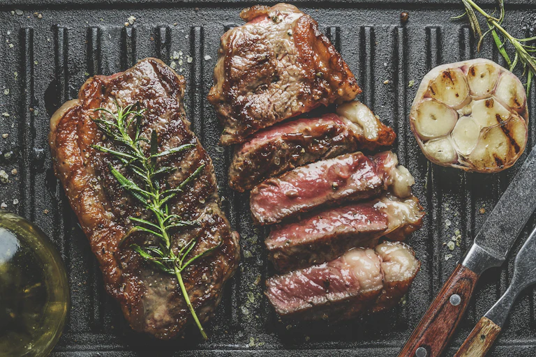 Barbecued steak with a rosemary sprig on top is cooking on a grill.
