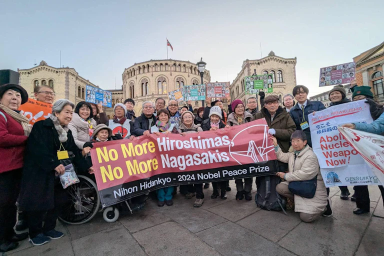 A group of people stand in a public square holding a banner reading 'No more Hiroshimas, no more Nagasakis.'