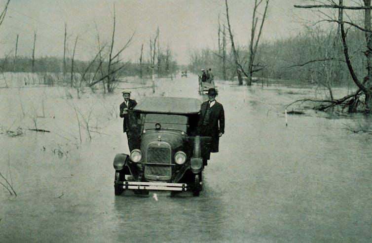 Three men ride on the outside of a car going through floodwater all around it.