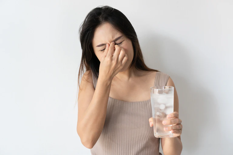 Une jeune femme tenant un verre d'eau glacée dans une main fait un geste de douleur.