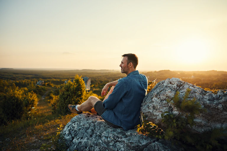A man sits on some rocks outdoors, watching the sunset.