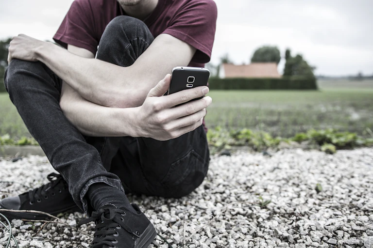 An anonymous young man or boy sitting on the ground in a rural area, holding his phone