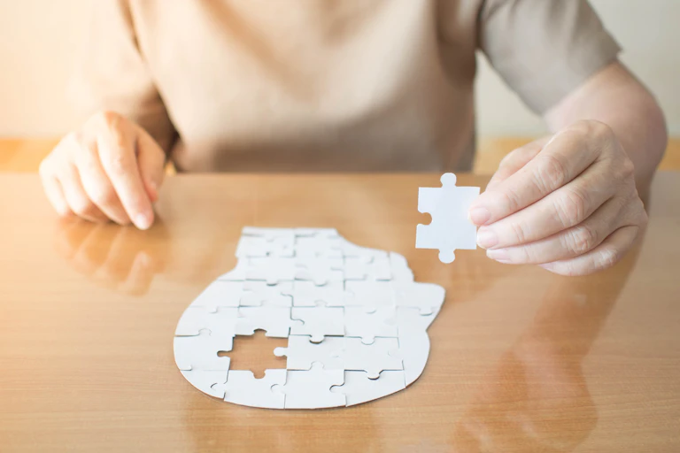 Woman's hands holding missing jigsaw puzzle piece. The unfinished jigsaw is the shape of a human head.