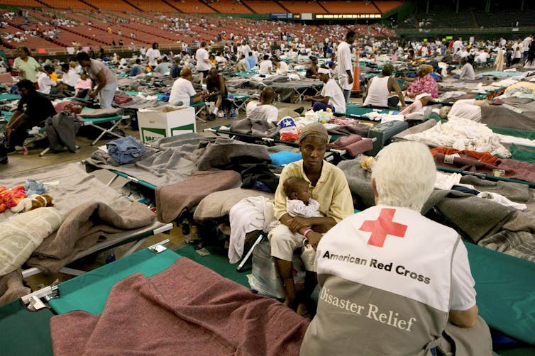 A woman with a Red Cross T-shirt talks to an evacuee holding a baby and sitting on a cot in the Superdome football stadium. The floor is filled with cots and people.