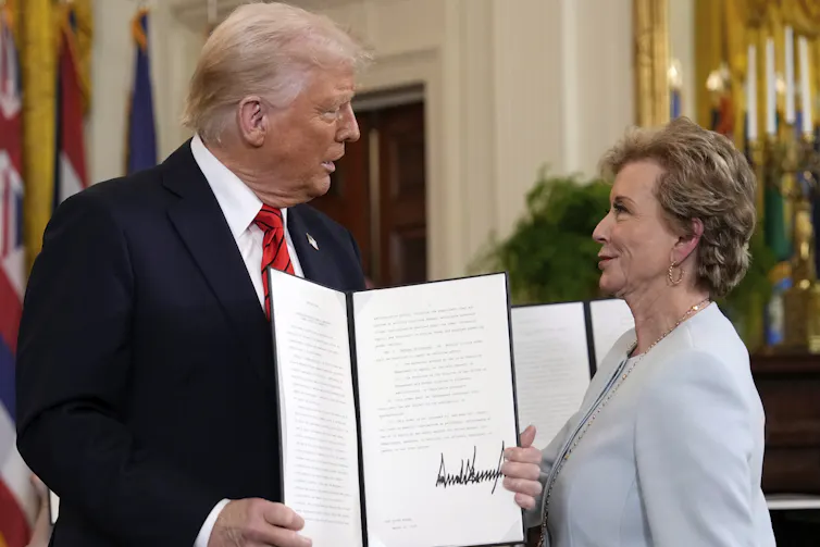An older man with white hair and a black blazer holds up a folder with paper inside it and faces an older woman wearing a light blue suit.