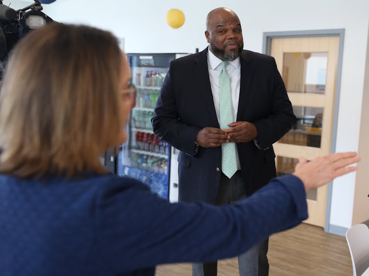 A middle-aged Black man wears a black blazer and white shirt and stands near a white woman with a navy blue blazer.