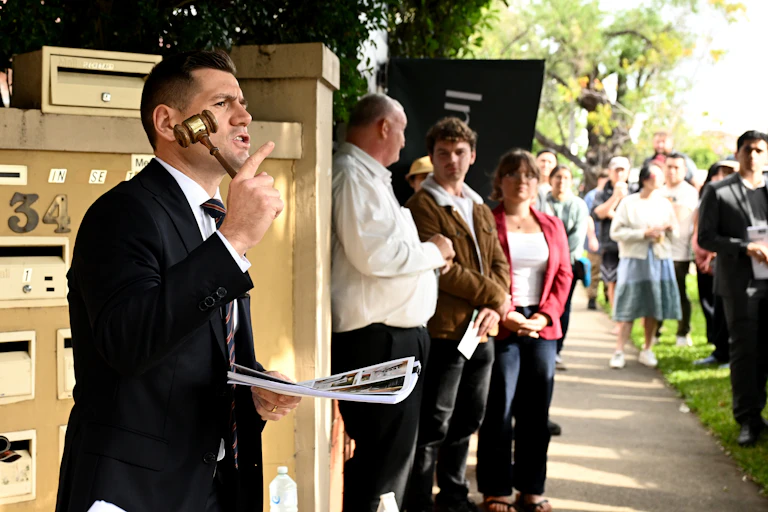 A male auctioneer holding a gavel, speaking to a large crowd at an auction in Sydney.