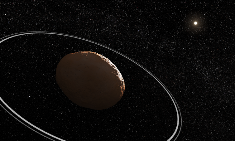 Një asteroid shkëmbor është në paradhomë dhe një pikë e ndritshme e mjegullt që përfaqëson Diellin është në sfond. Asteroidi ka dy unaza të ngushta rreth tij. Sfondi është i zi dhe plot yje.