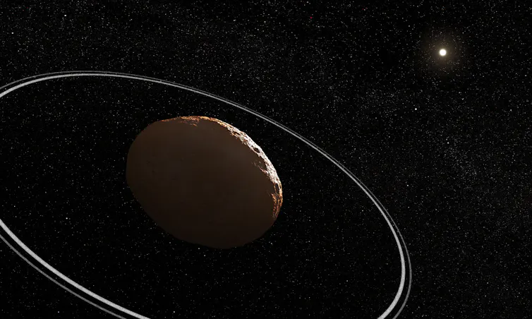 Um asteroide rochoso está em primeiro plano e um ponto brilhante e difuso representando o Sol está ao fundo. O asteroide tem dois anéis estreitos ao seu redor. O fundo é preto e cheio de estrelas.