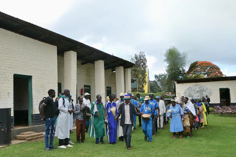 Men and women clapping and playing drums on a grassy patch of land with low buildings around them