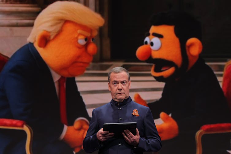 Dmitry Medvedev delivers a speech in front of caricatures of Trump and Zelensky.