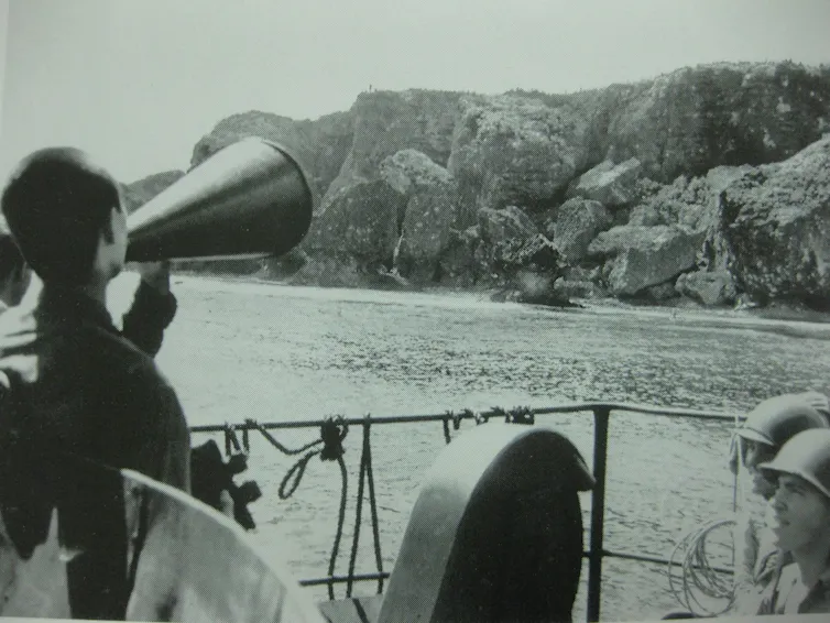 Archival photograph of a Japanese POW shouting through a loudhailer from a ship to an island.