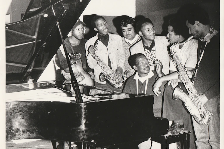 An old black and white photo of young to middle-aged African men, one seated at a paino and surrounded by the others, some holding musical instruments.