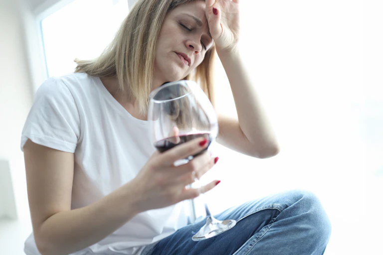Une femme se tient la tête, avec un verre de vin rouge à la main.