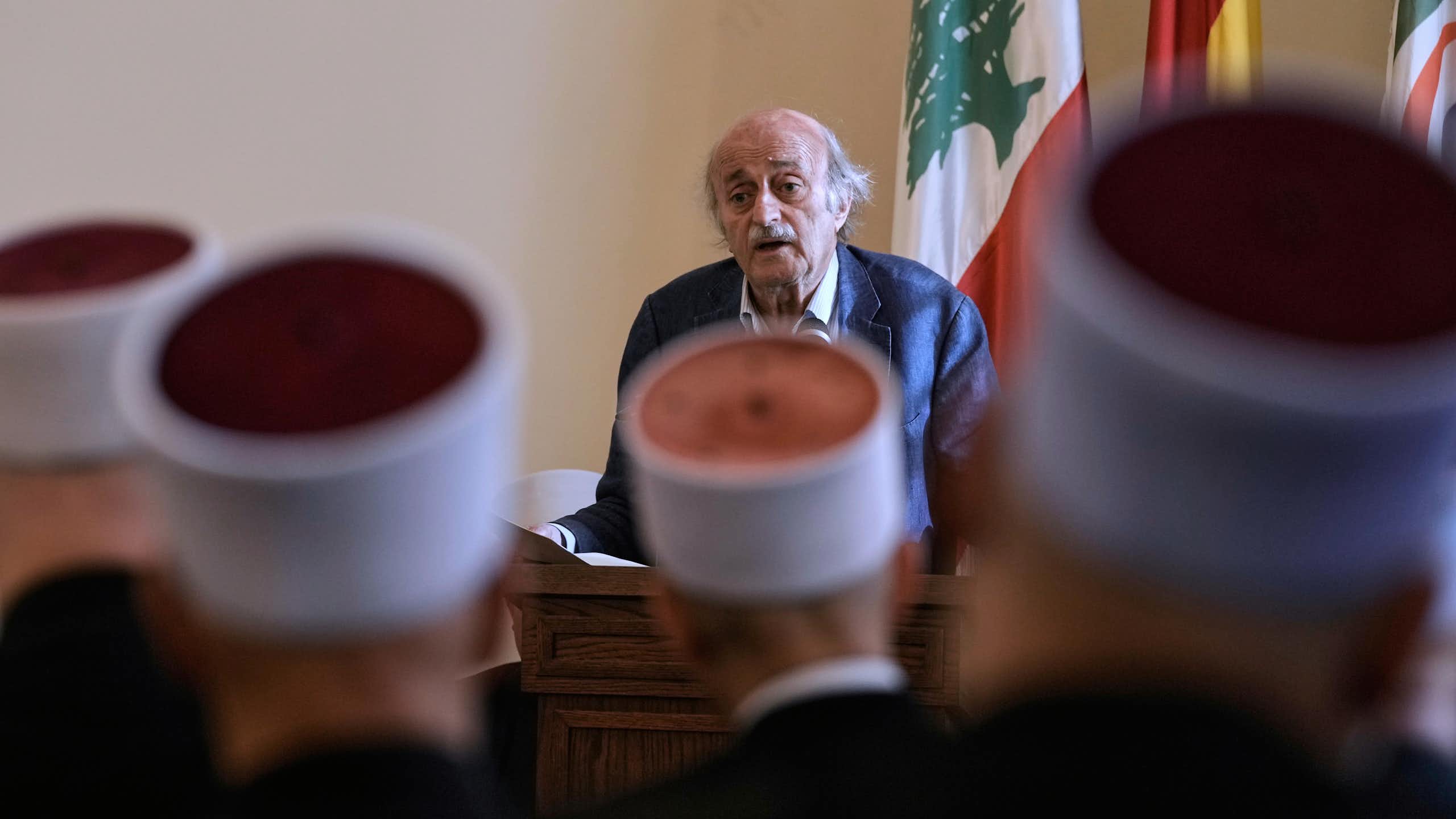 An elderly man talks to people wearing round hats who are seated in front of him.