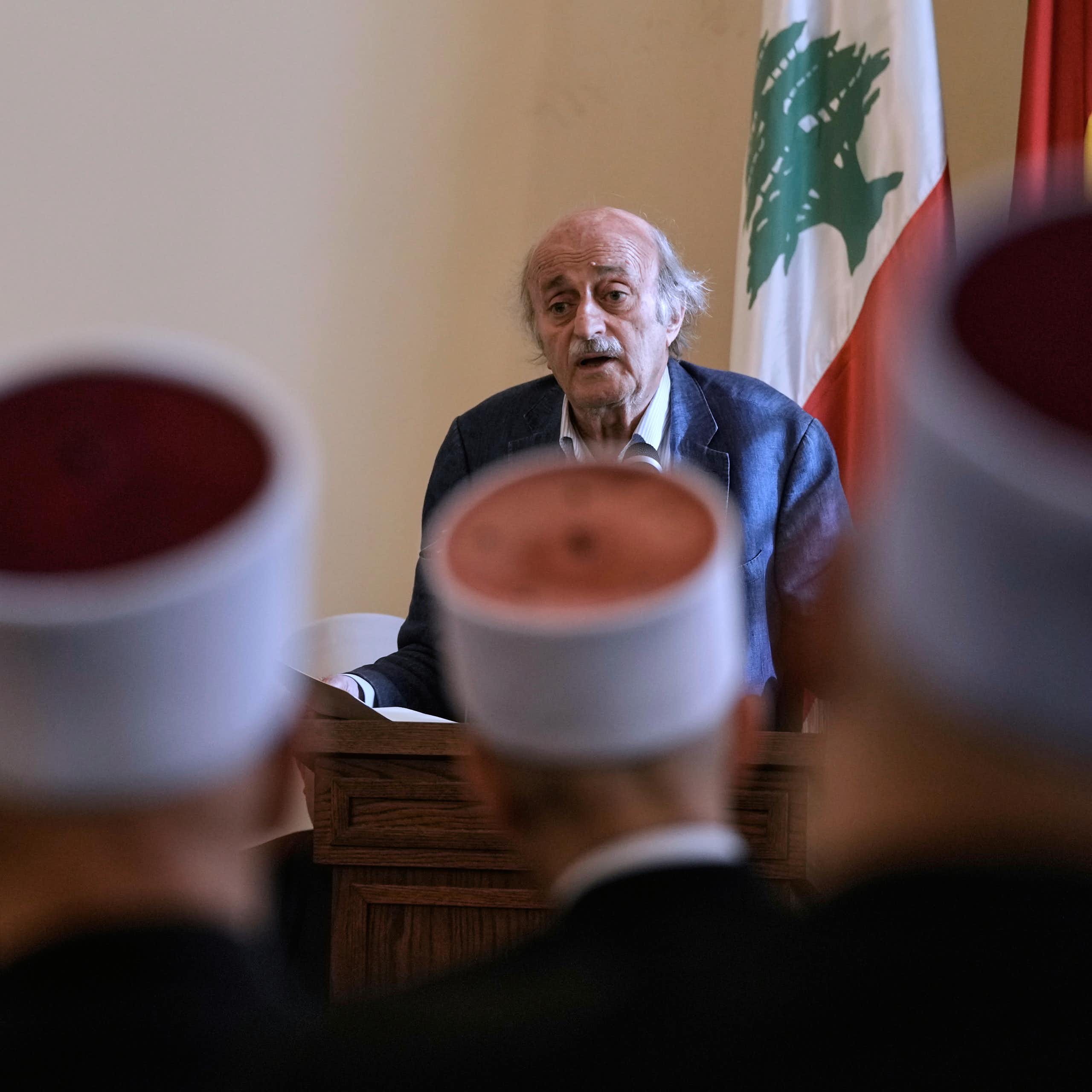 An elderly man talks to people wearing round hats who are seated in front of him.