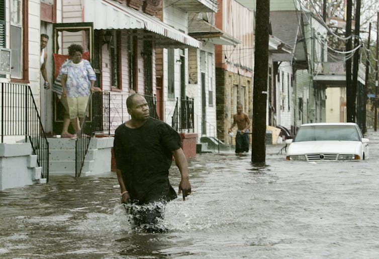 A man with a destressed look walks in thigh-deep water while people watch from a doorway several steps above street level.
