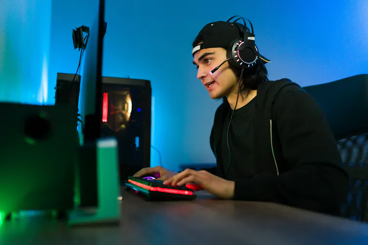 El joven estĆ” sentado frente al escritorio con un mouse y un teclado. Ćl usa auriculares