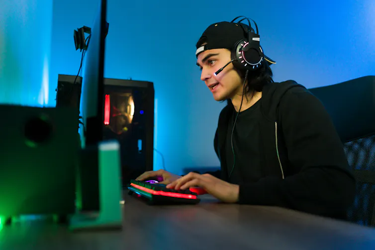 A young man sits in front of a desktop computer using a mouse and keyboard. He is wearing headphones